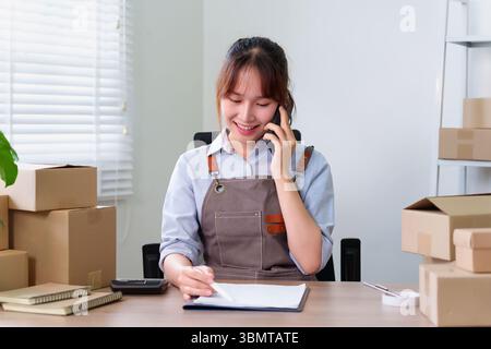 Young adult Asian woman entrepreneur calling on smartphone checking order with delivery boxes. Small SME startup e-commerce packaging products from ho Stock Photo