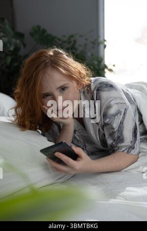 A young woman is enjoying a blissful moment in bed, smiling with her smartphone, surrounded by greenery and light Stock Photo