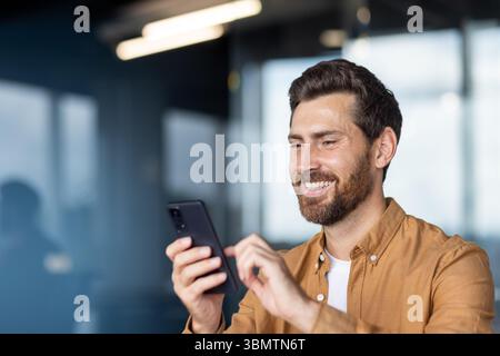 A smiling man in a button-down shirt looks at his phone, likely texting or browsing in a modern office setting. Stock Photo