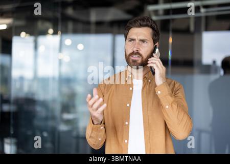 A concerned businessman communicates on his smartphone, gesturing animatedly while looking upwards in an office setting. Stock Photo