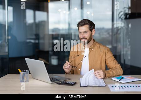 A businessman in a modern office setting reviews documents while using a laptop. He is holding his glasses. Stock Photo
