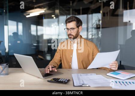 A businessman at his office desk, working on a laptop, holding papers, analyzing data, and wearing glasses. Stock Photo