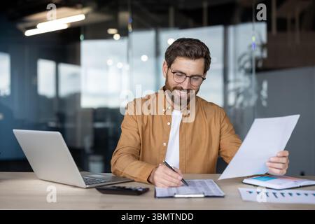 A cheerful businessman with glasses reviews financial documents in his office, using a laptop and calculator for analysis. Stock Photo