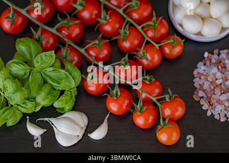 Fresh cherry tomatoes on vine with basil leaves, garlic cloves, Himalayan pink salt and mozzarella balls on black wooden table Stock Photo