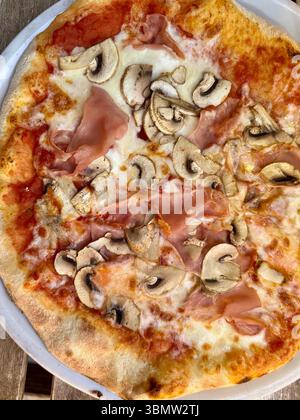 Close-up of a traditional Italian pizza with mozzarella cheese, ham, tomato sauce, and fresh mushrooms on a thin crust, served on a white plate. Stock Photo