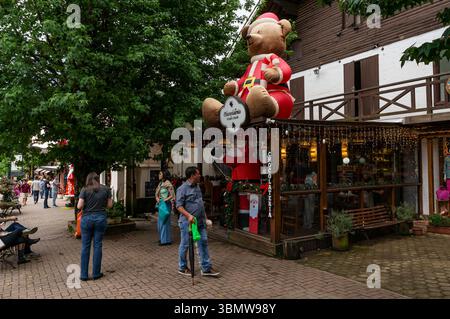 People walk past Chocolateria Monte Verde shop featuring giant inflatable Santa bear on roof, string lights, and tree at main avenue. Stock Photo