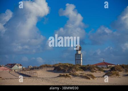 A tall, white clock tower with a distinct top stands prominently amidst sandy dunes and scattered buildings under a bright blue sky filled with dramat Stock Photo