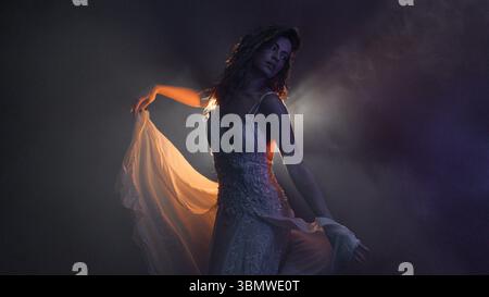 Graceful dancer in flowing white gown, silhouetted by mysterious backlight in moody atmosphere Stock Photo