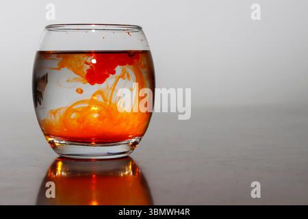 Closeup Image of Orange Ink Swirl in Glass of Water on a Reflected Wooden Table in White Background Stock Photo