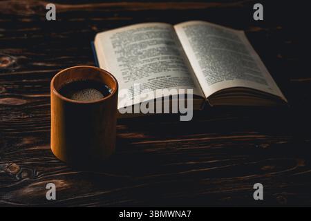 A ceramic coffee cup sits beside an open book on a dark wooden table, lit by warm light, evoking a peaceful reading moment Stock Photo
