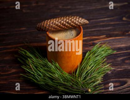 A ceramic cup of coffee sits next to a pine cone and evergreen branch on a dark wooden table, evoking a warm and rustic forest-inspired setting Stock Photo