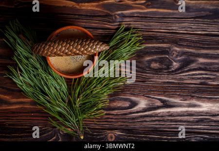 A ceramic cup of coffee sits next to a pine cone and evergreen branch on a dark wooden table, evoking a warm and rustic forest-inspired setting Stock Photo