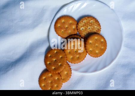Golden round crackers are stacked on a white plate placed on soft white fabric, captured in natural daylight with a simple, clean composition Stock Photo
