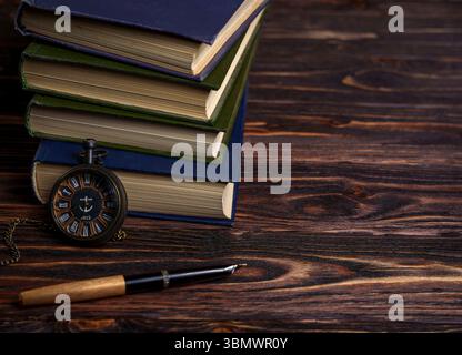 A stack of books is placed on a rustic wooden table alongside a vintage pocket watch and fountain pen, evoking a scholarly and timeless atmosphere Stock Photo