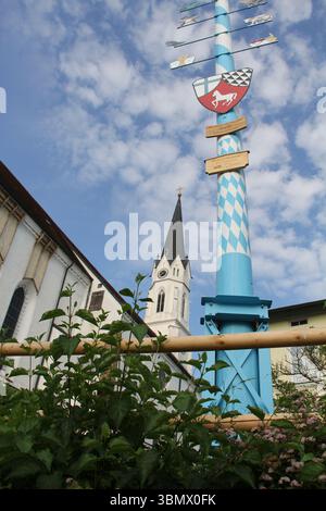 KIRCHDORF, GERMANY - JUNE 19, 2025: Upward view of traditional blue-and-white maypole and church steeple in Kirchdorf framed by plants against a Stock Photo