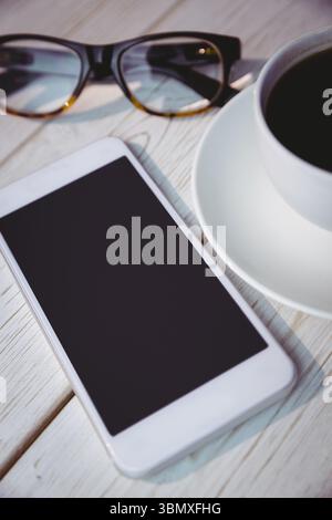 Showing pale wooden tabletop featuring white smartphone, black-framed eyeglasses and coffee cup Stock Photo
