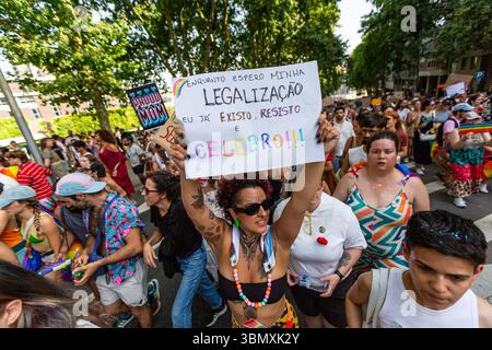 A Pride marcher holds a hand-painted sign that reads “Enquanto espero minha legalização eu já existo, resisto e celebro!!!” (“While I wait for my legalization, I already exist, resist, and celebrate!!!”) during the 20th LGBTI+ Pride March. Surrounded by a lively, colorful crowd—including signs of family support and vibrant outfits—the image captures the intersection of identity, resilience, and joyous protest in the streets of Porto on June 28, 2025, Portugal. Stock Photo