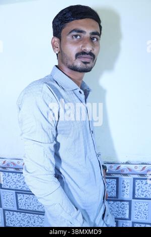 Portrait of a young Indian man standing indoors against a plain wall, wearing a light grey shirt Stock Photo