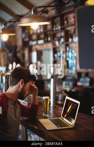 Laptop sitting on wooden bar counter beside pint of beer under industrial pendant lights in pub Stock Photo