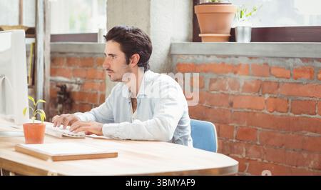 Man typing on keyboard at wooden desk in loft office beside computer monitor, notebooks and plants Stock Photo