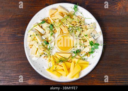 An artistic arrangement of various cheeses alongside honey, nuts, and fresh microgreens is presented on a white plate. The rustic wooden table adds wa Stock Photo