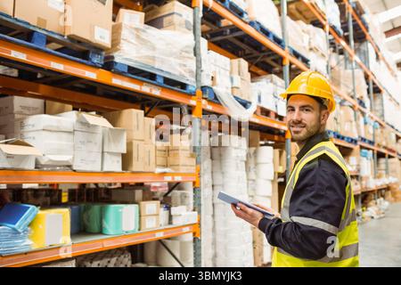 Male worker wearing hard hat and hi-vis vest inspecting stock with tablet in warehouse, copy space Stock Photo