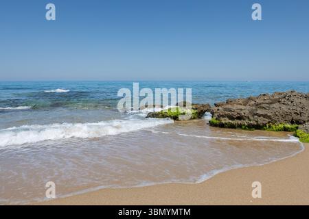 Waves hitting algae-covered rocks on a sunny coastal beach with clear turquoise water Stock Photo