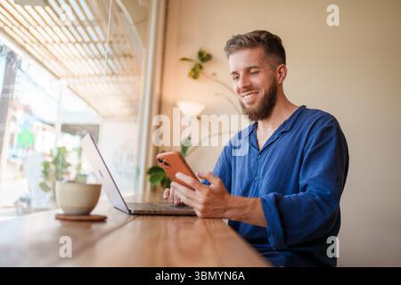 A Smiling Young Man is Clearly Focused on His Laptop While Working in a Cozy Cafe Environment Stock Photo