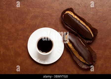 coffee, chocolate Eclair, coffee in a white Cup, white saucer, on a brown table, Eclair on paper stand Stock Photo