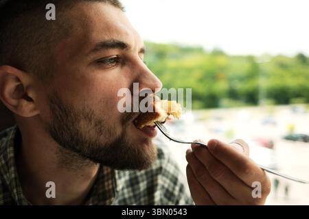 Bearded man about to eat a bite of blueberry cake, holding fork near his mouth and looking ahead in a bright, natural-lit cafe. Close side view portra Stock Photo