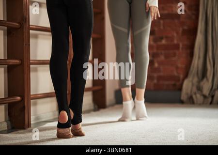 Two Caucasian teenagers practicing rhythmic gymnastics training, standing on toes in ballet position, lower bodies visible, focusing on technique and posture in gym environment Stock Photo