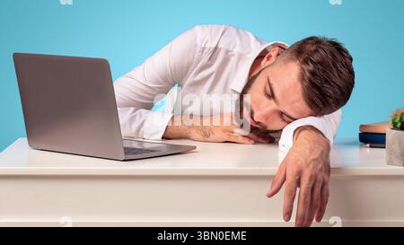 Exhausted young businessman sleeping on his desk, tired of overworking on blue studio background, copy space Stock Photo