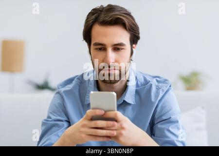 Serious Man Using Smartphone Wearing Earbuds Sitting On Sofa Indoors Stock Photo