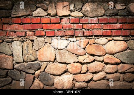 A textured wall displays a blend of smooth stones and red bricks positioned evenly. Soft natural light highlights the materials' colors and shapes, cr Stock Photo