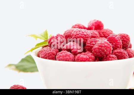 A close-up shot of ripe fresh raspberries in a white bowl with green leaves on a light background. Concept of healthy eating, summer harvest, and orga Stock Photo