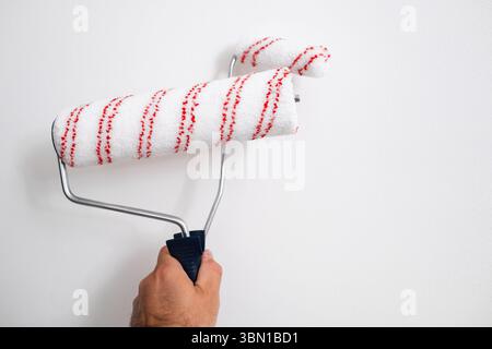Male hand holding a large and small paint rollers while painting a white wall, close up of home renovation process.  Stock Photo