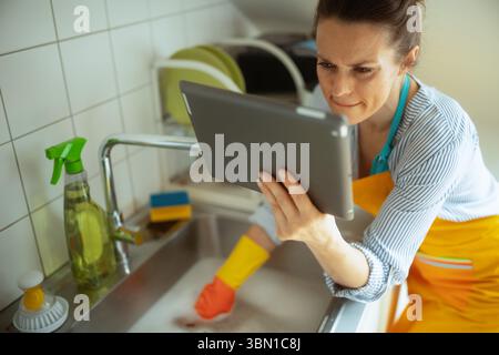 A woman in gloves and apron uses a tablet while facing a clogged kitchen sink, searching for help or instructions to resolve a drain issue. Stock Photo