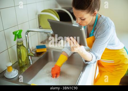 Woman in orange apron uses a tablet while cleaning a soapy stainless steel sink with gloves and cleaning supplies in a modern kitchen. Stock Photo