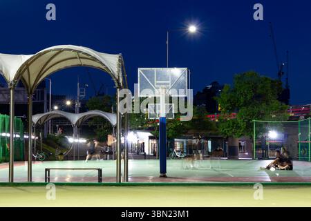 Night scene captures a public basketball court illuminated by bright lights, with blurred figures engaged in activity, set against a deep blue sky Stock Photo