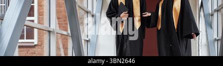 Header of two anonymous young students wearing graduation gowns standing in hallway talking and holding diploma folders, university students engaging in conversation after ceremony Stock Photo
