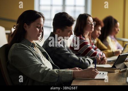 Polyethnic group of university students sitting in classroom taking notes and using laptop, young adults and teenagers focusing on studying during lecture, side view of diverse group Stock Photo