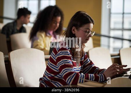 Caucasian young woman sitting in lecture hall using digital tablet, multiethnic university students studying in background, focused on academic work, casual classroom setting Stock Photo