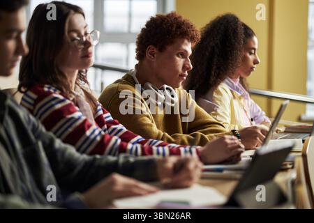 Diverse group of university students, including Caucasian and Black teenagers, sitting at desks, listening attentively and taking notes, during classroom lecture with laptops and notebooks Stock Photo