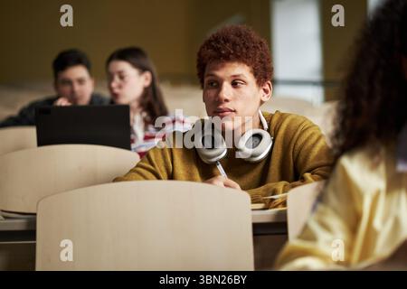 Young Black man sitting in lecture hall wearing headphones around neck, writing notes while listening to lecture, other university students studying in background Stock Photo