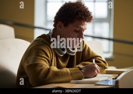 Teenage Black man sitting at desk writing in notebook with over ear headphones around neck in university classroom setting, focused on studying and taking notes during lecture Stock Photo
