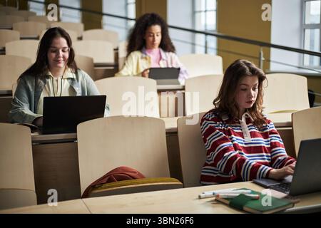 Three young female students sitting in university lecture hall using laptops, focusing on studying and taking notes, multiethnic group engaged in academic work during class session Stock Photo