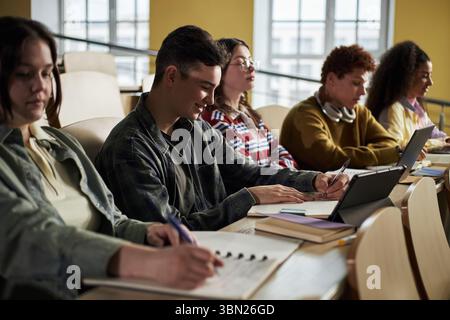 Group of diverse university students sitting in lecture hall taking notes and using digital tablets, young people including Caucasian and Black individuals focusing on academic work during class Stock Photo