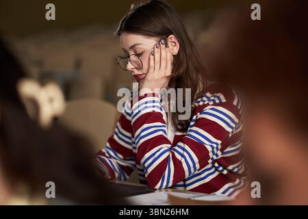 Caucasian young woman wearing glasses, sitting at desk, studying in university classroom, resting head on hand, appearing focused on academic work, surrounded by blurred students Stock Photo