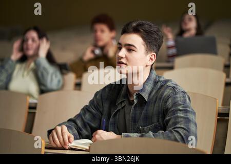Caucasian young man sitting in lecture hall taking notes in notebook while listening attentively, with multiethnic university students blurred in background using laptops and talking Stock Photo