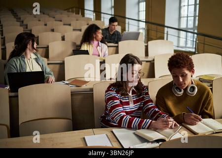 Group of multiethnic university students sitting in lecture hall working on assignments, young women and men collaborating with notebooks and laptops during study session Stock Photo